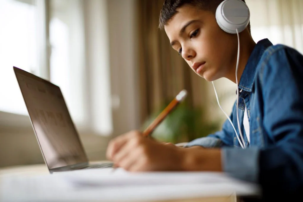 Youth with headphones writing in notebook in front of a laptop.