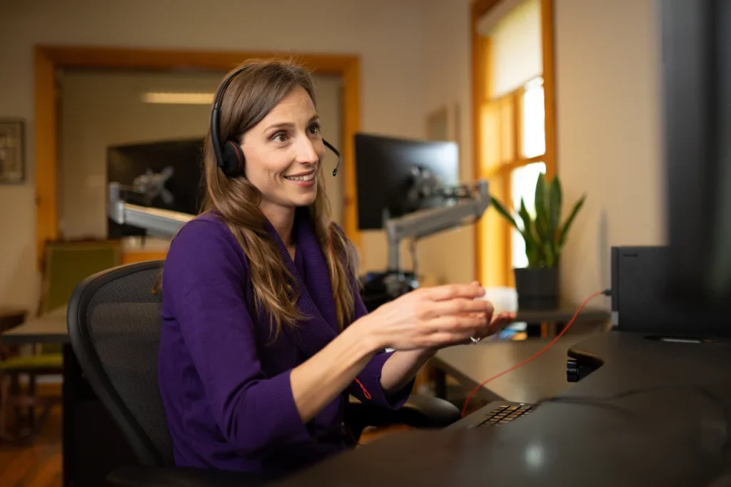 A teacher wearing a headset in front of a computer providing online tutoring support to a student.
