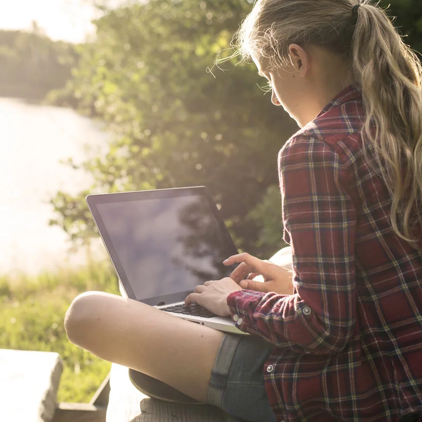 A girl sitting on a bench with a laptop in front of a lake.
