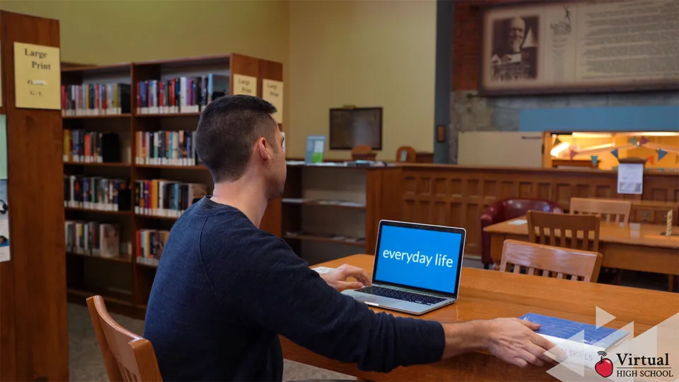 A man sitting at a table in a library with a laptop.