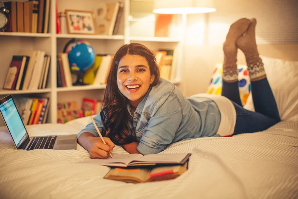 A student laying on her bed with a laptop and notebook.